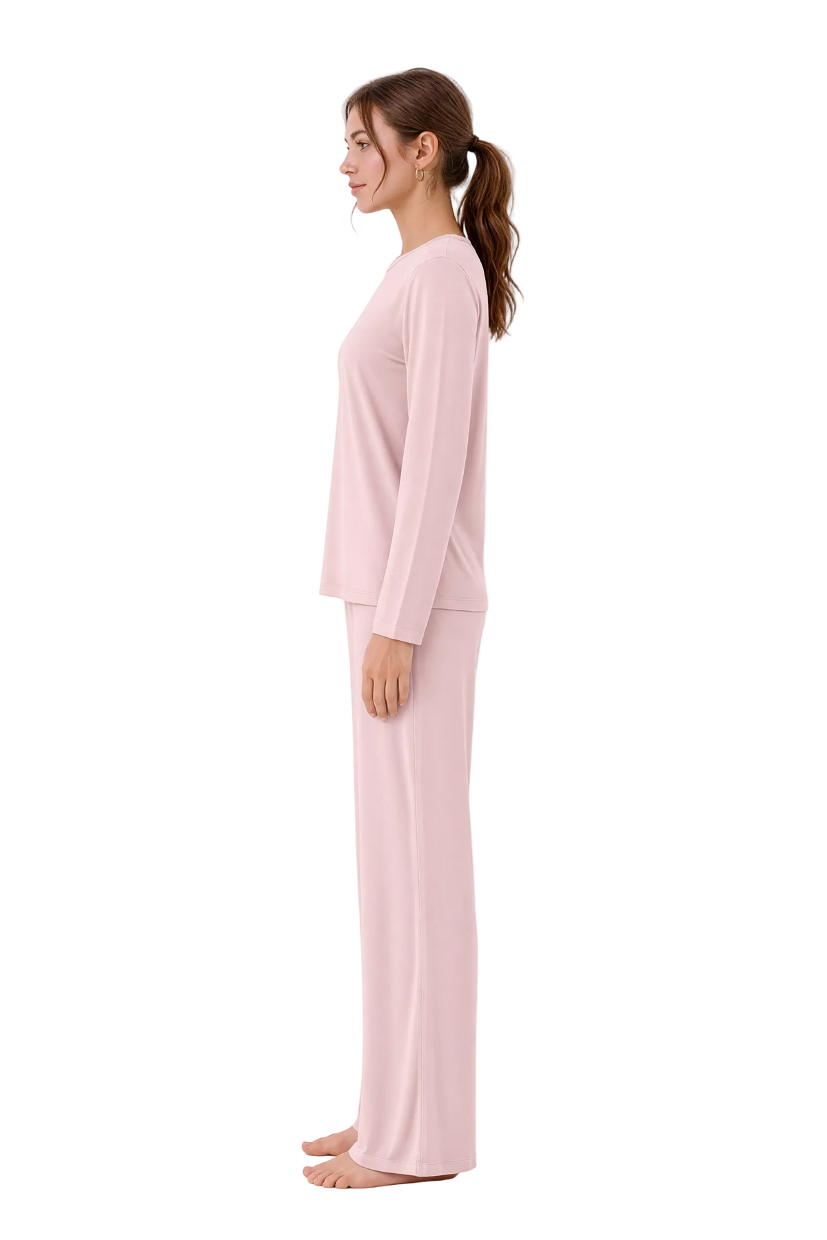 A woman stands in profile wearing a light pink two-piece pajama set with a long-sleeve top and matching pants against a plain white studio background.