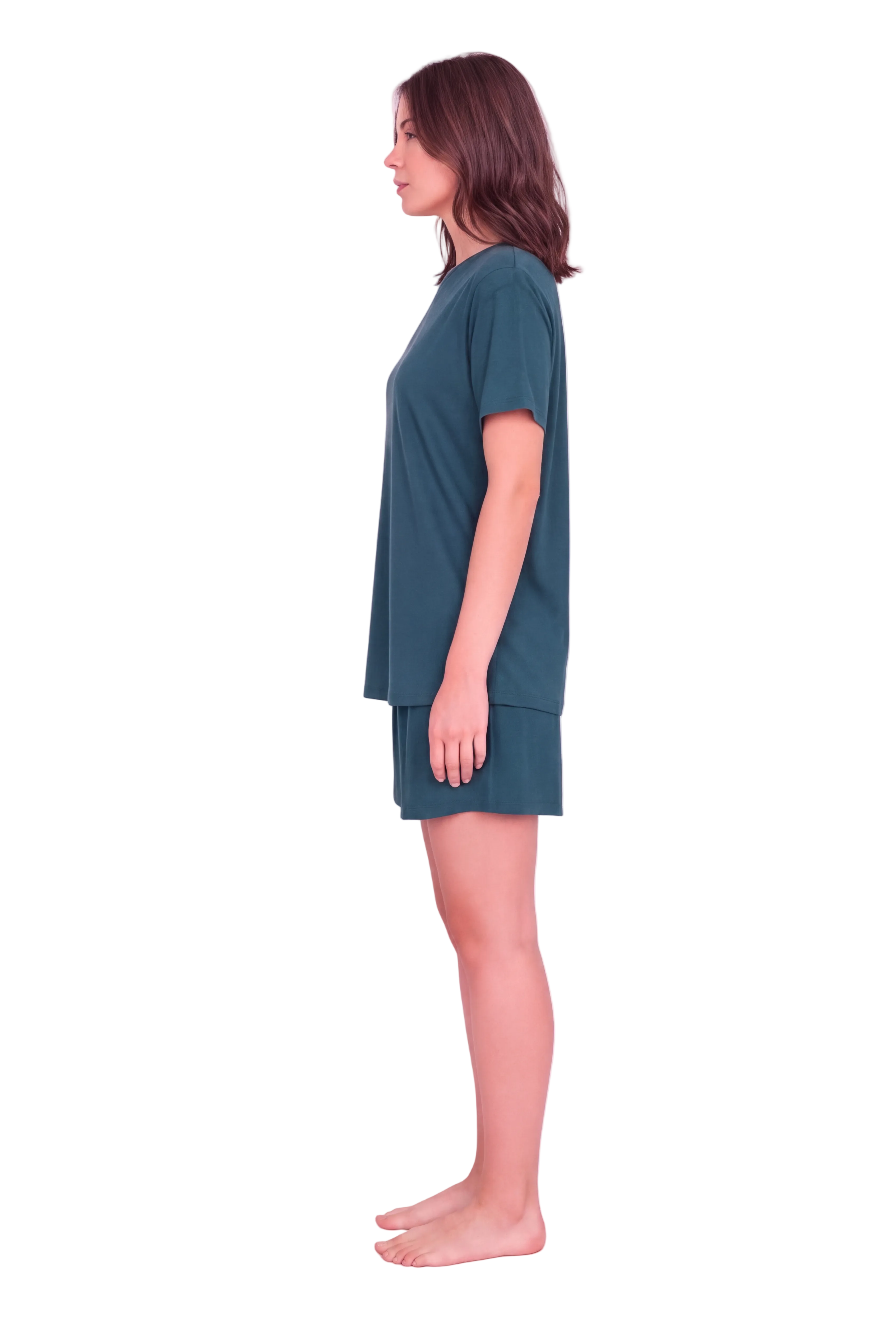 A woman stands in profile wearing a matching two-piece lounge set consisting of a short-sleeve top and shorts in a muted teal green color, set against a solid pale pink studio background.